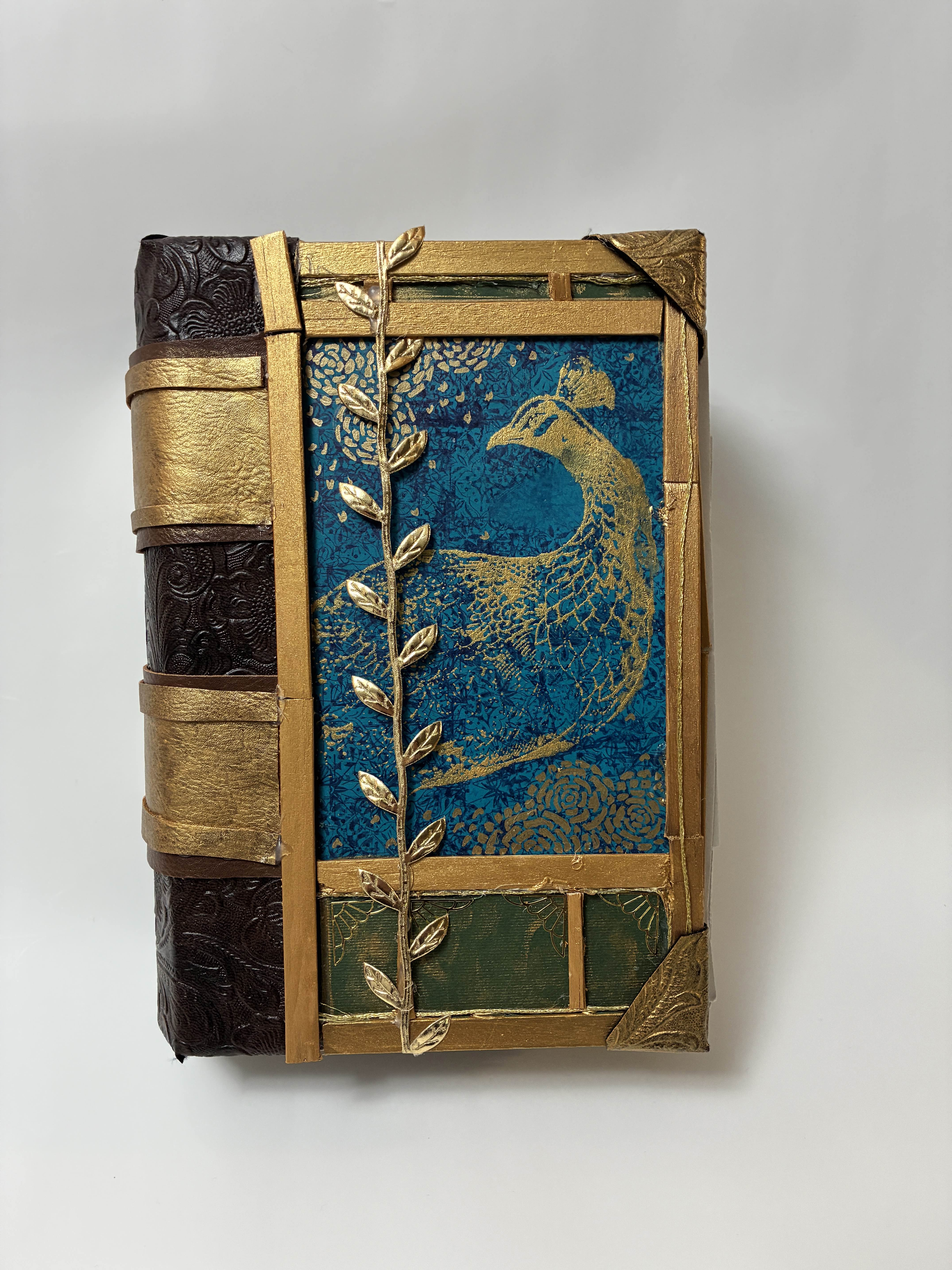 photograph of the final product (the hallow book box) on a white background. The box itself is deep brown letter spine adored with golden straps, the cover is framed by gold, accented with gold ribbon of leaves, the top and bottom are separate into frames of green, and the center of the cover is dark blue with a gold painting of a peacock