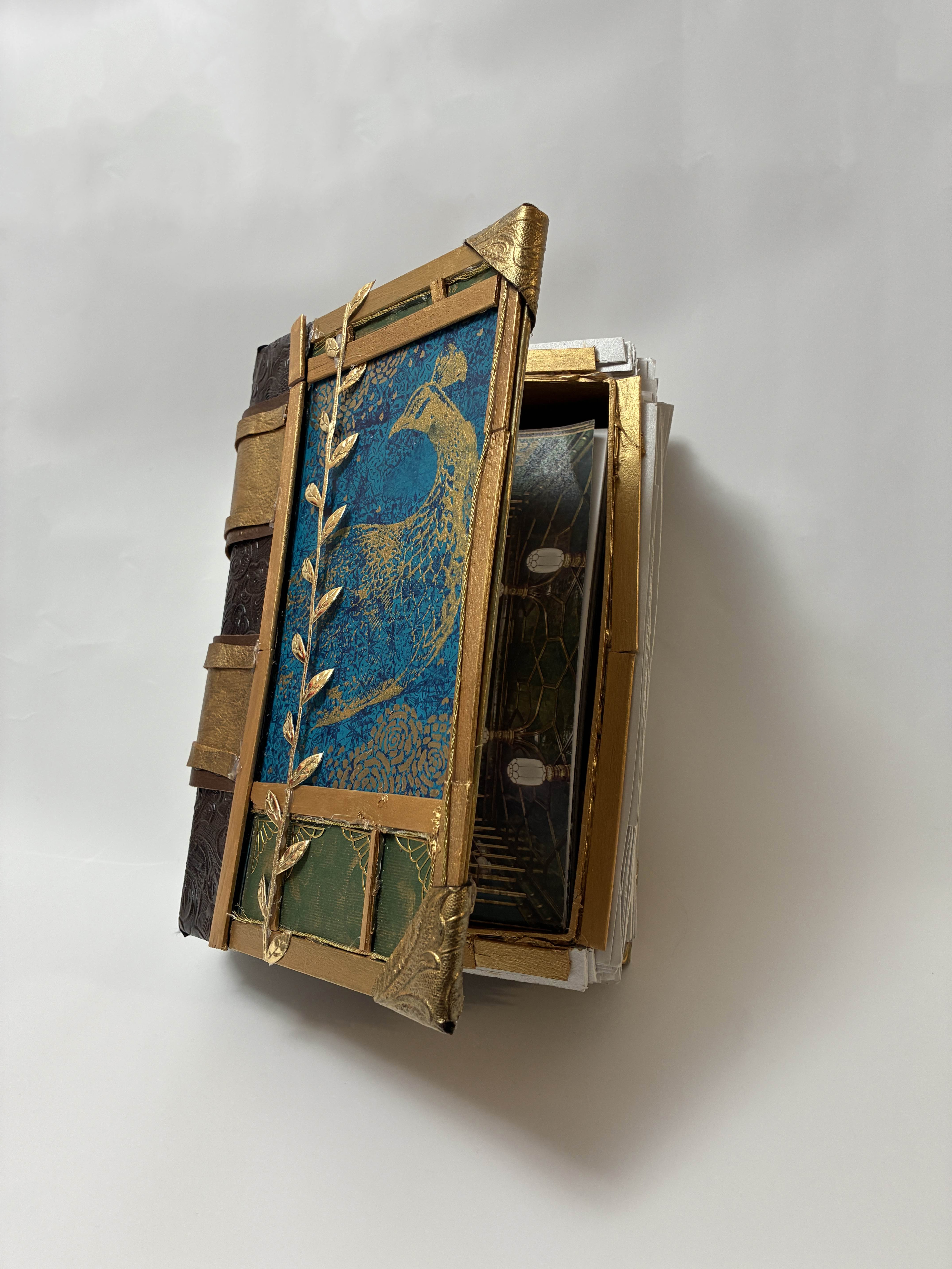 photograph of the final product (the hallow book box) on a white background with the cover opened, so the viewer can see the objects contained inside. The box itself is deep brown letter spine adored with golden straps, the cover is framed by gold, accented with gold ribbon of leaves, the top and bottom are separate into frames of green, and the center of the cover is dark blue with a gold painting of a peacock